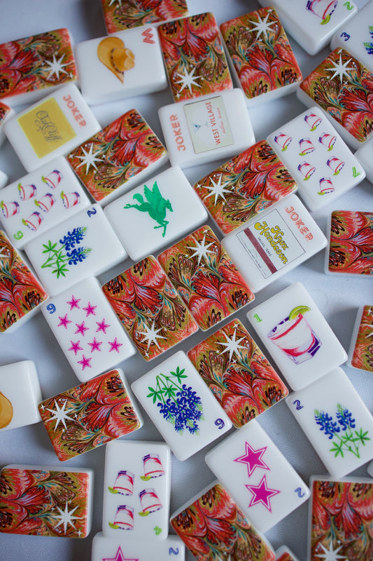 Dallas Collective mahjong tiles scattered on a white tablecloth, with bluebonnet, pink star, cocktail and Joker tiles among marbled red-and-gold tile backs